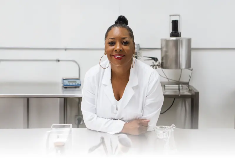 Paula Hayes, founder of Hue Noir, in a lab coat, smiling in her cosmetics lab, championing beauty for people of color.