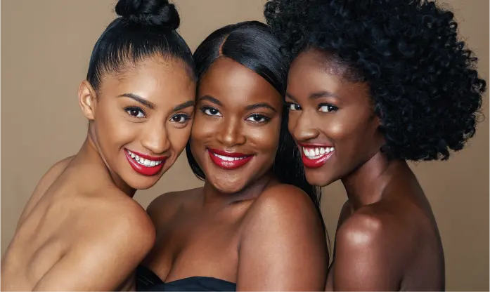 Three radiant women with diverse skin tones, glowing complexions, and bold red lipstick pose closely against a warm backdrop.