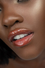 Model with medium deep skin tone wearing coral red lipstick and shimmery clear gloss overlay